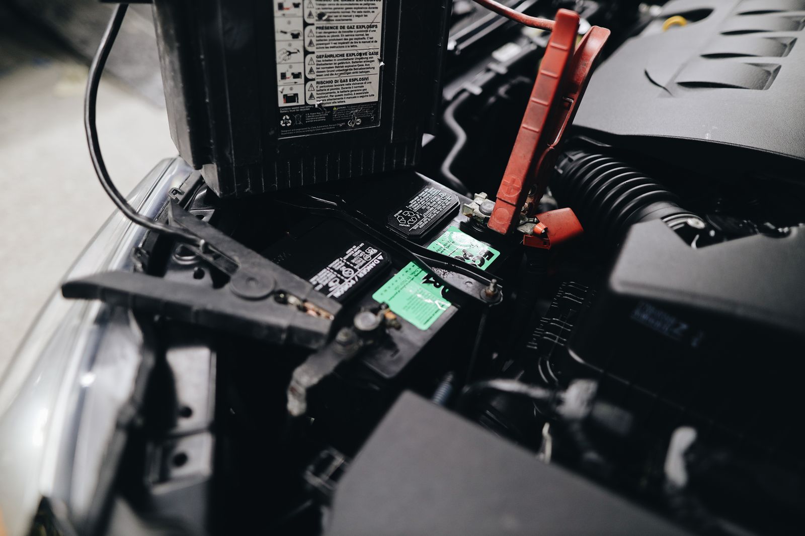 A car battery in an engine bay with a red jumper cable clamp on the positive terminal and a printed explosive-gas warning label on the case